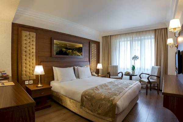 Innenansicht eines Doppelzimmers im Yigitalp Hotel mit einem Doppelbett, Nachttischen, Stühle und ein Fenster mit Vorhängen.