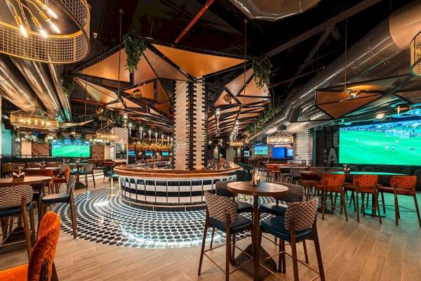 Innenbereich der Bar im FIVE LUXE JBR Hotel mit Tischen, Stühlen und einer großen Anzeigetafel.