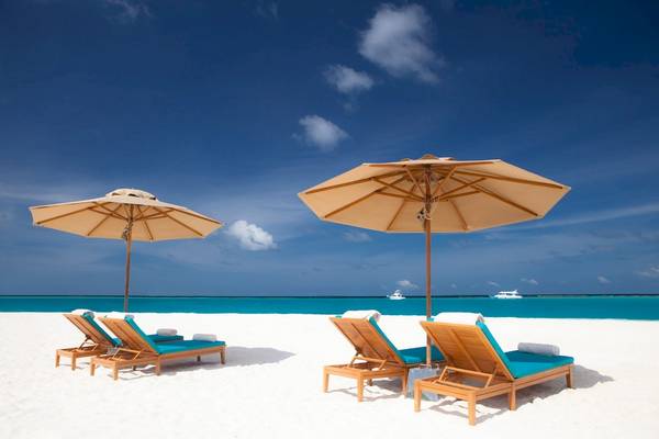 Drei Sonnenliegen mit blauen Kissen und beigefarbenen Sonnenschirmen auf einem weißen Sandstrand.