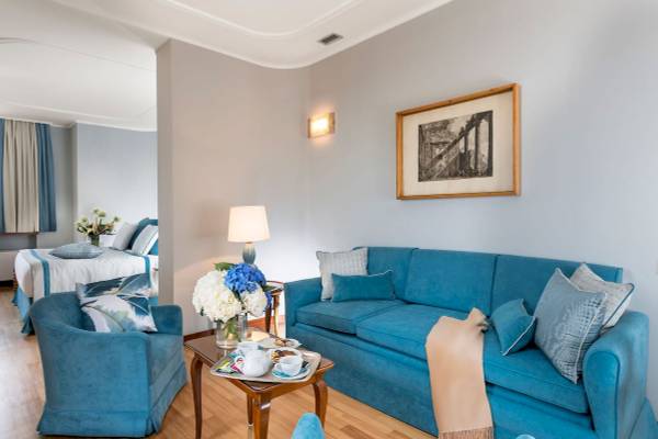 Innenansicht der Suite im Bettoja Hotel Mediterraneo mit blauem Sofa, Tisch, Blumenvase und Bild an der Wand.