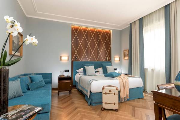 Zimmer im Bettoja Hotel Mediterraneo mit einem Bett, Sofa, Vase mit Blumen und einem Koffer.
