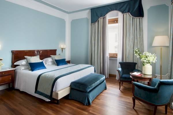Schlafzimmer im Bettoja Hotel Mediterraneo mit einem großen Bett, blauen Kissen, und einem blumigen Fenstervorhang.