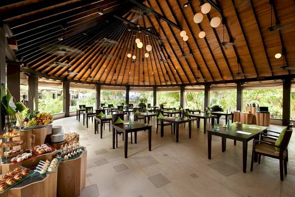 Innenbereich des Sun Siyam Iru Fushi Restaurants mit runden Tischen und Stühlen, tropischen Dekorationen und einer Auswahl an Speisen.
