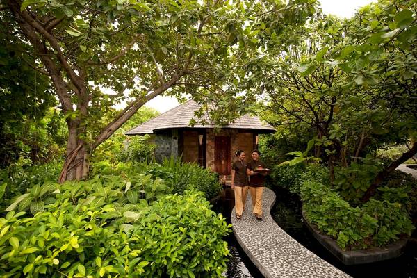 Zwei Frauen tragen ein Tablett durch einen gepflasterten Weg im Freien des Sun Siyam Iru Fushi Hotels.