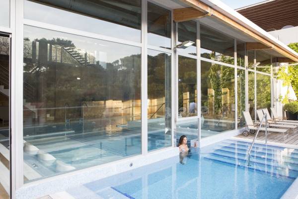 Eine Frau schwimmt im blauen Pool des ARTIEM Audax Hotels mit großen Glaswänden und Sonnenliegen draußen.