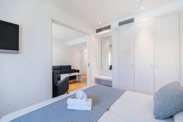 Schlafzimmer mit Bett und Couch im Hotel Princesa Playa, weiße Wände und Möbel, Fernseher an der Wand, Spiegel, offene Tür zum Wohnzimmer.