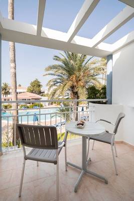 Hotelbalkon mit Aussicht auf Pool und Palmen, zwei Stühle und ein Tisch mit süßen Leckereien.