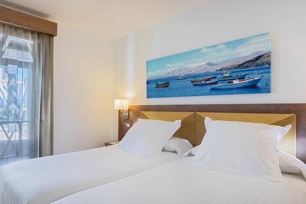 Doppelzimmer mit Meerblick im Barcelo Fuerteventura Castillo Hotel mit zwei Betten, weißem Dekor und einem Gemälde von Booten auf See.