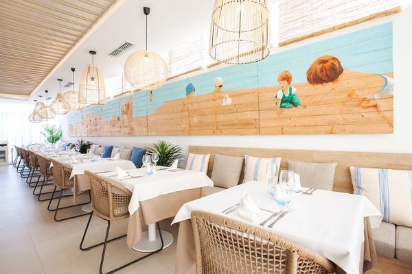 Innenansicht des Restaurants des Grupotel Aldea Cala'n Bosch mit gemauerten Wänden, Tischdekoration und einem großen Wandbild von Kindern am Strand.
