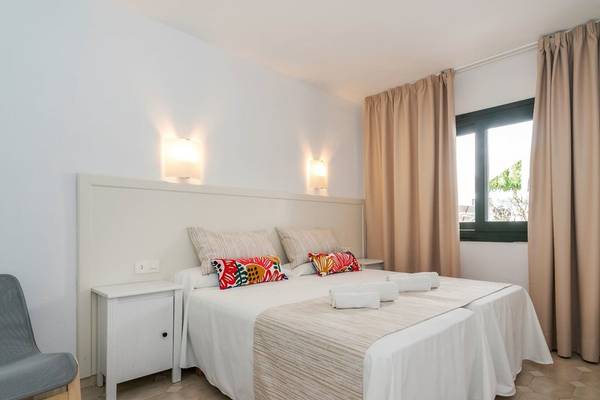 Schlafzimmer mit Doppelbett im Beach Club Menorca, weiße Wände, Bettwäsche und Nachttische, bunte Kissen, beleuchtete Wandlampen.