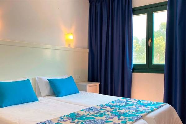 Schlafzimmer im Beach Club Menorca mit zwei Betten, blauen Kissen, weißen Decken und blauen Vorhängen.