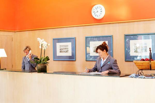 Rezeption im Grupotel Playa Club mit zwei Angestellten, einer schreibt und die andere telefoniert, und Dekorelementen im Hintergrund.