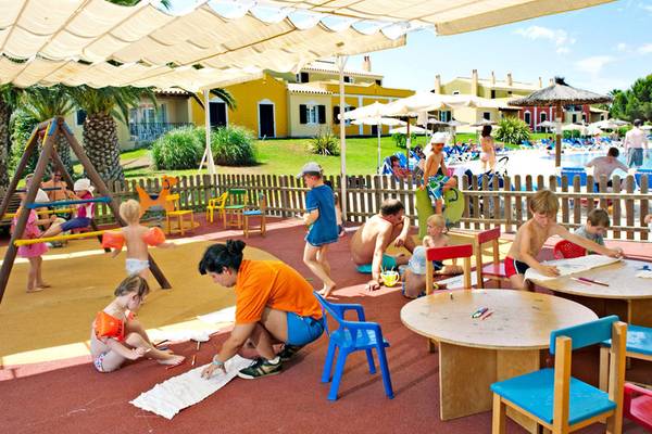 Kinder spielen auf einem Spielplatz im Grupotel Playa Club, umgeben von blauen Stühlen und Tischen.