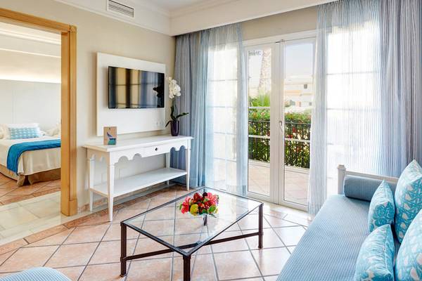 Innenansicht einer Suite im Grupotel Playa Club Hotel mit Wohnzimmer, Couch, Tisch und Schlafzimmer im Hintergrund.