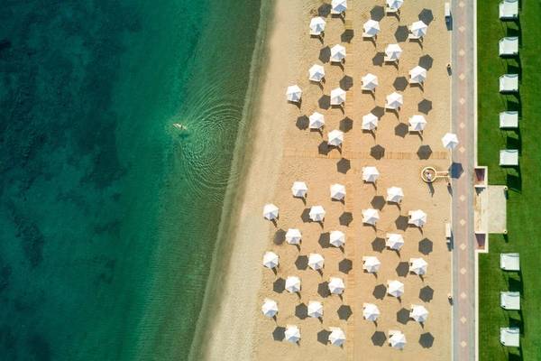 Luftaufnahme des Miraggio Thermal Spa Resorts Strands mit vielen weißen Sonnenschirmen und einer Person im Wasser.