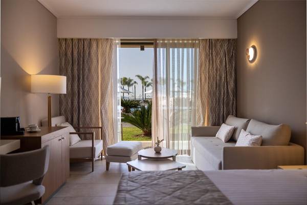 Innenansicht eines Zimmers im Miraggio Thermal Spa Resort mit Schlafbereich, Wohnzimmer, Couch, Tisch, Lampen und Blick auf Palmen.