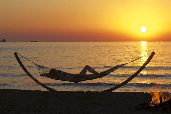 Person entspannt sich in einer Hängematte bei Sonnenuntergang am Meer.
