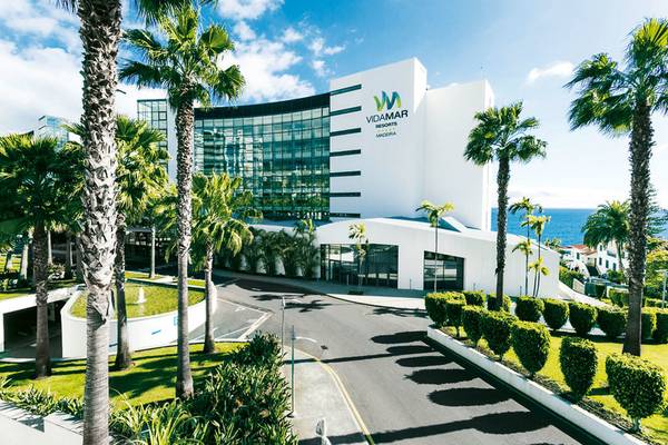 Außenansicht des VidaMar Resort Hotel Madeira mit weißen Gebäuden, Glasfassaden und Palmen im Vordergrund.