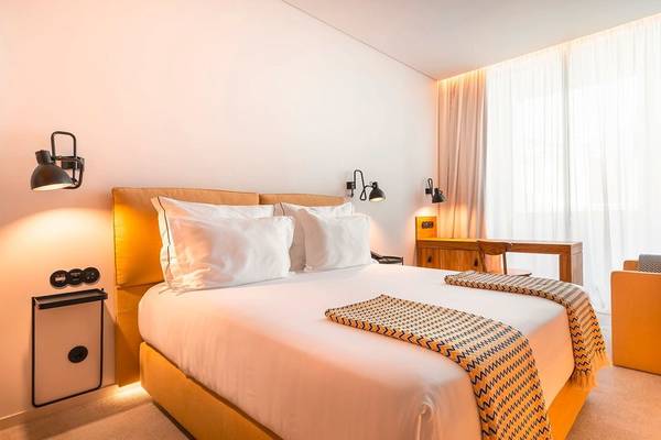 Innenansicht eines Hotelzimmers im 3HB Faro mit einem Bett, Kissen, Decken, Wandleuchten und einem Schreibtisch.