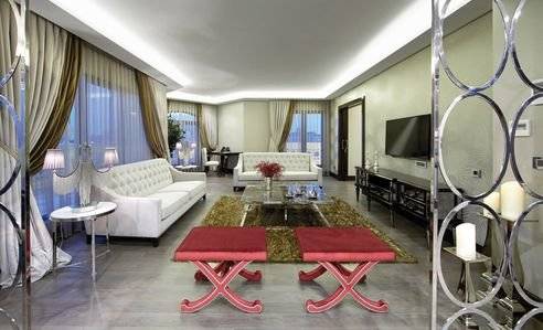 Wohnzimmer im DoubleTree by Hilton Istanbul Esentepe mit weißen Möbeln, einem Fernseher und rot gepolsterten Hockern.
