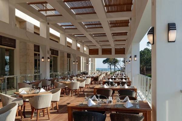 Innenansicht des Restaurants im Rixos Premium Belek Hotel mit Holztischen und Stühlen auf einer Terrasse mit Meerblick.