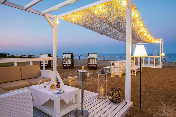 Loungebereich des Asterias Beach Resorts am Abend mit beleuchteten Überdachungen und Möbeln auf dem Strand.