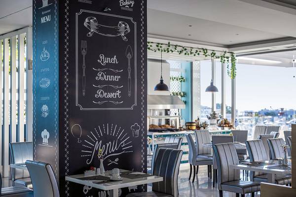 Innenbereich des Asterias Beach Resort Restaurants mit einem großen schwarzen Menüboard, das neben einem Tisch steht, mit Blick auf einen sonnigen Hafen.
