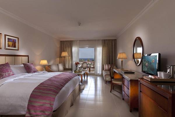 Innenansicht eines komfortablen Hotelzimmers im Baron Palace Sahl Hasheesh mit Doppelbett, Fernseher, Schreibtisch und Balkon.