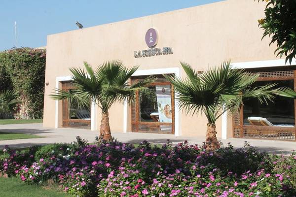 Eingang zur La Siesta Spa im Gemma Resort mit zwei Palmen und Blumen im Vordergrund.