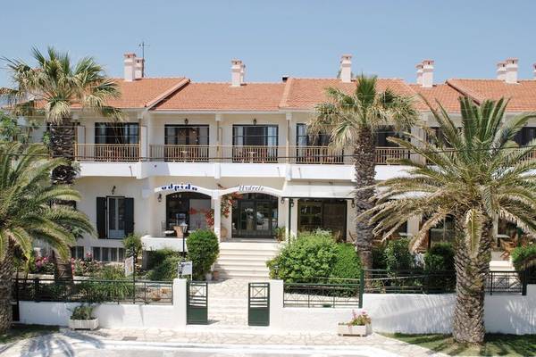 Front view of the Hydrele Beach Hotel with red roofs and white walls surrounded by palm trees and bushes.