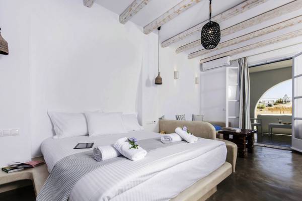 Innenansicht eines Zimmers im Cycladic Islands Hotel & Spa mit einem großen Bett, weißen Kissen und Decken, Holzbalken an der Decke und einer offenen Tür.