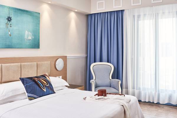 Hotelzimmer im Lagos Mare mit Bett, Kissen, Stuhl, Fenstervorhang und Gemälde an der Wand.