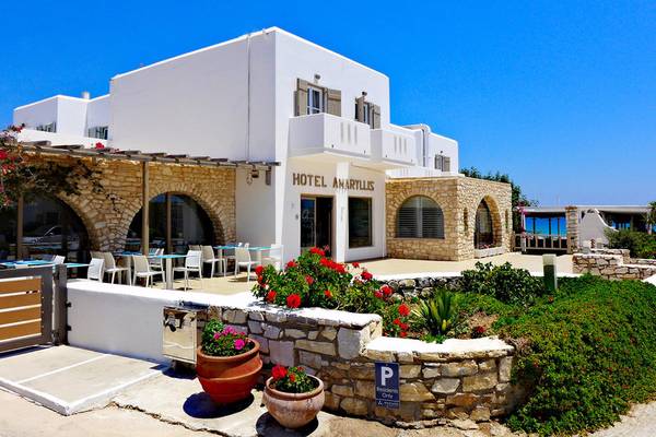 Außenansicht des Amaryllis Paros Beach Hotels mit weißem Gebäude, Balkonen, Tisch und Stühlen, roten Blumen und Parkplatzschild.