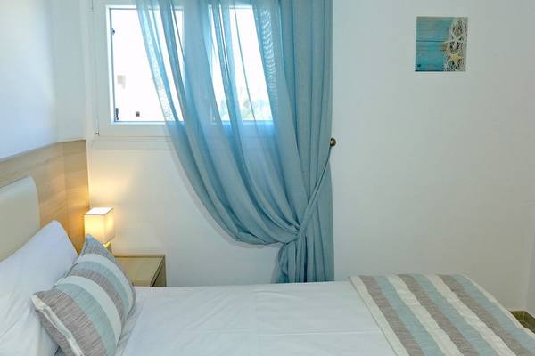 Ein modernes Hotelzimmer mit einem Bett, einem Nachttisch, einer Lampe und einem Fenster mit einem Vorhang im Amaryllis Paros Beach Hotel.