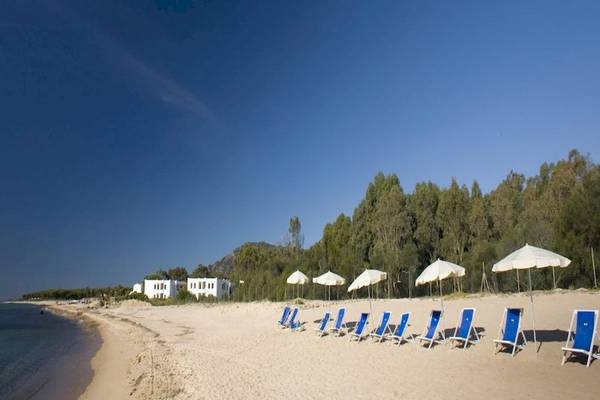 Ein Strand mit blauen Liegen und weißen Sonnenschirmen, hinter denen das Mare Pineta Hotel steht.