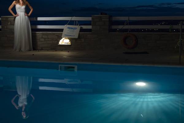 Eine Frau im weißen Kleid steht am Rand eines Swimmingpools bei Nacht.