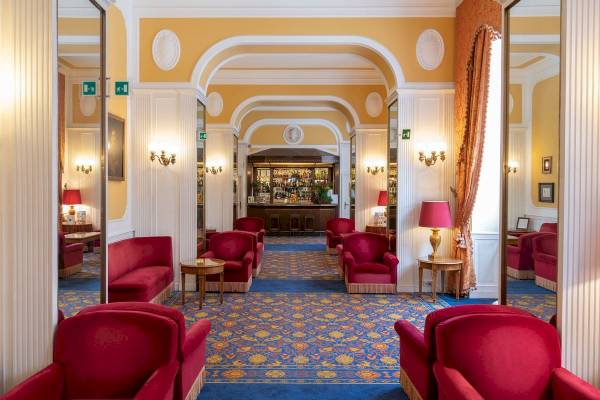 Der geräumige und luxuriöse Loungebereich des Bettoja Hotel Massimo d'Azeglio mit roten Sesseln, blauem Teppich und einer Bar im Hintergrund.
