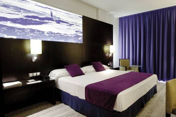Innenansicht eines Hotelzimmers im Vincci Maritimo mit Doppelbett, Nachttischen und blauen Vorhängen.