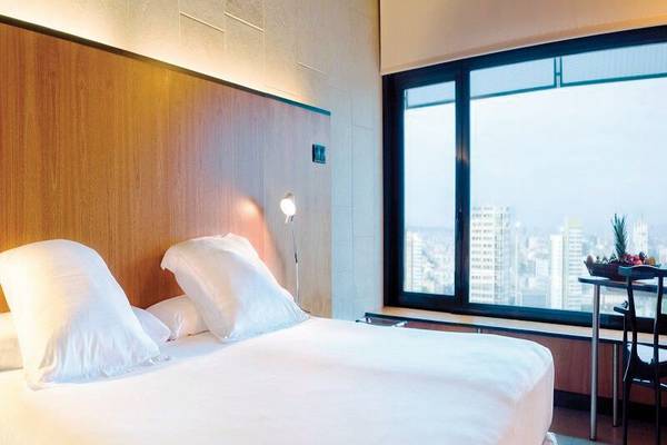 Ein modernes Hotelzimmer mit einem Bett, weißen Kissen, einer Lampe und einem Fenster mit Stadtblick im Hintergrund.