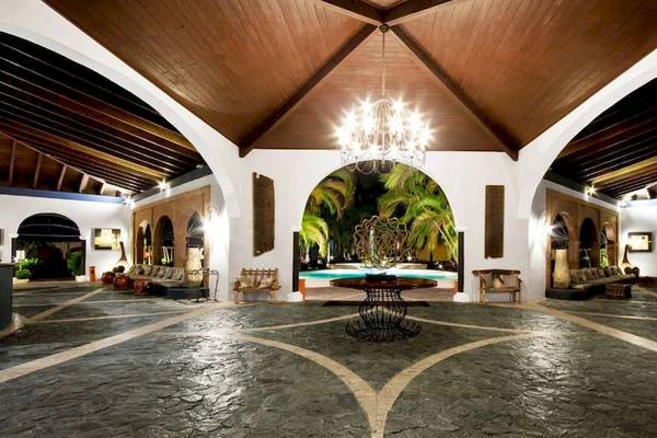Innenansicht des Hotel-Lobbys des Catalonia Bayahibe mit einer großen Deckenleuchte, einem zentralen Tisch, einem Swimmingpool im Hintergrund und Möbeln.