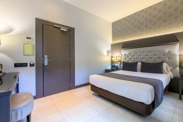 Innenansicht eines Hotelzimmers im Ciutadella Barcelona mit Doppelbett, Nachttischen und grauem Stuhl.