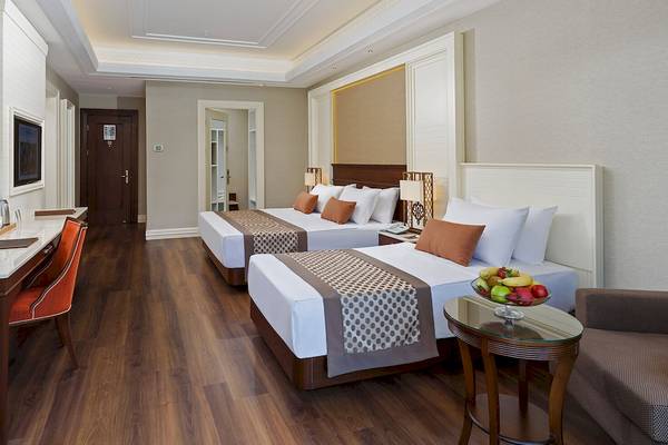 Innenansicht eines Zimmers im Güral Premier Belek Hotel mit zwei Betten, Schreibtisch und Früchtekorb auf einem Tisch.