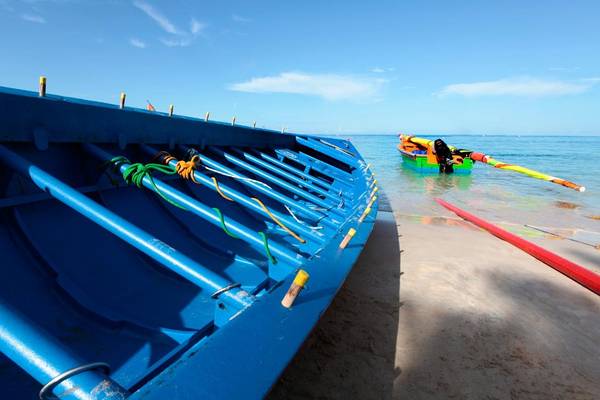 Ein blaues Boot am Strand mit einem farbenfrohen Ruderboot im Hintergrund.