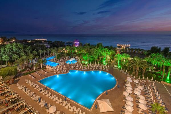 Abendliche Außenansicht des Delphin Deluxe Hotels mit großen Swimmingpools und beleuchteten Palmen.