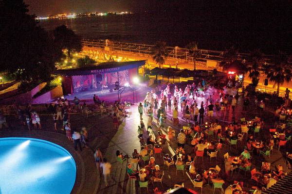 Abendveranstaltung im Melas Holiday Village mit Pool, Bühne und vielen Gästen bei Nacht.