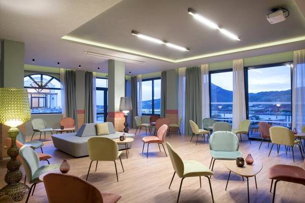 Hotellobby des Dolce Athens Attica Riviera mit farbenfrohen Stühlen und Tischen, großen Fenstern und Aussicht auf die Berge.