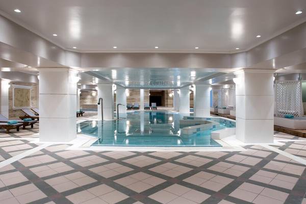 Innenansicht des Schwimmbads im Dolce Athens Attica Riviera Hotel mit weißen Säulen und gefliestem Boden.