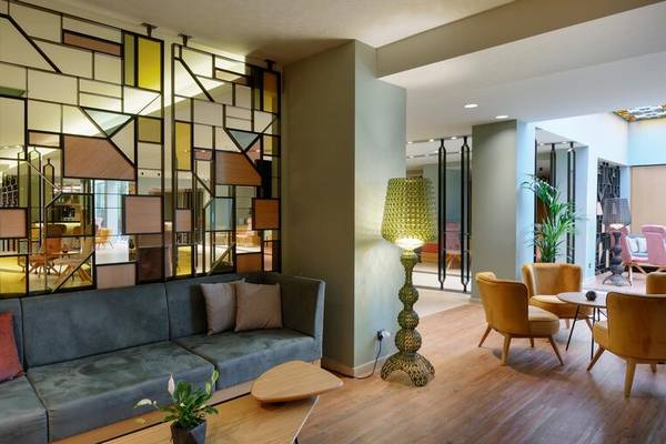 Sitzecke im Hotellobby von Dolce Athens Attica Riviera mit einem grauen Sofa, Kissen, und einer gelben Stehlampe.