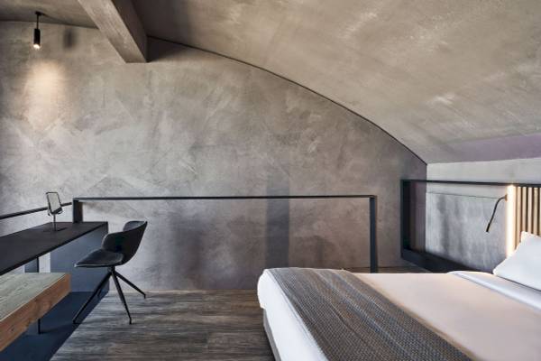Innenansicht des Schlafzimmers im Nema Design Hotel mit Betonwand, Schreibtisch, Stuhl und Bett.