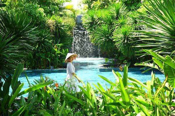 Eine Frau im weißen Kleid und Hut steht im TIA Wellness Resort Garten neben einem Pool und Wasserfall.